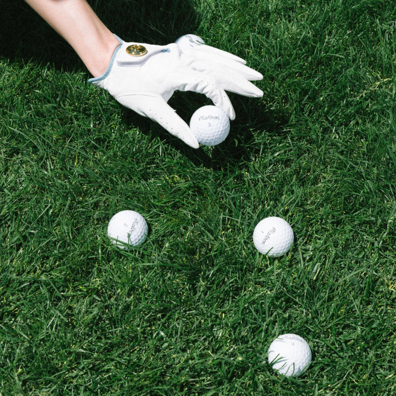 Golf balls on grass with a St John Knits Malbon gloved hand reaching towards one of them